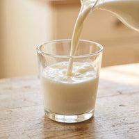 Milk being poured from a white pitcher into a clear glass on a wooden table.