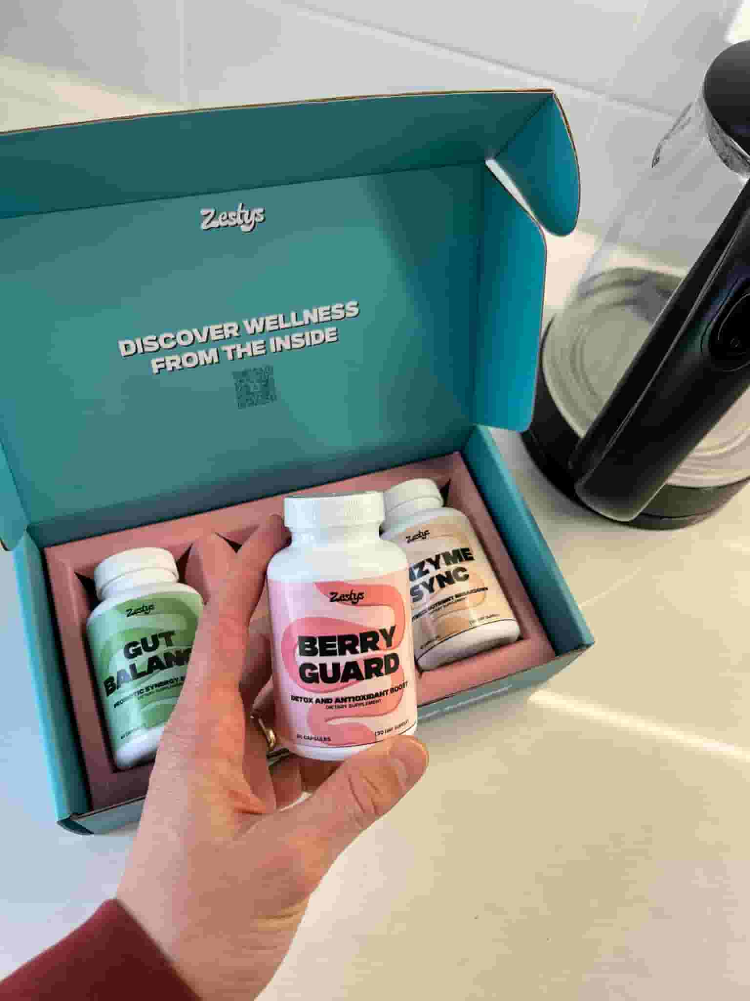 Hand holding a supplement bottle labeled 'Berry Guard' in front of an open box with more supplements.